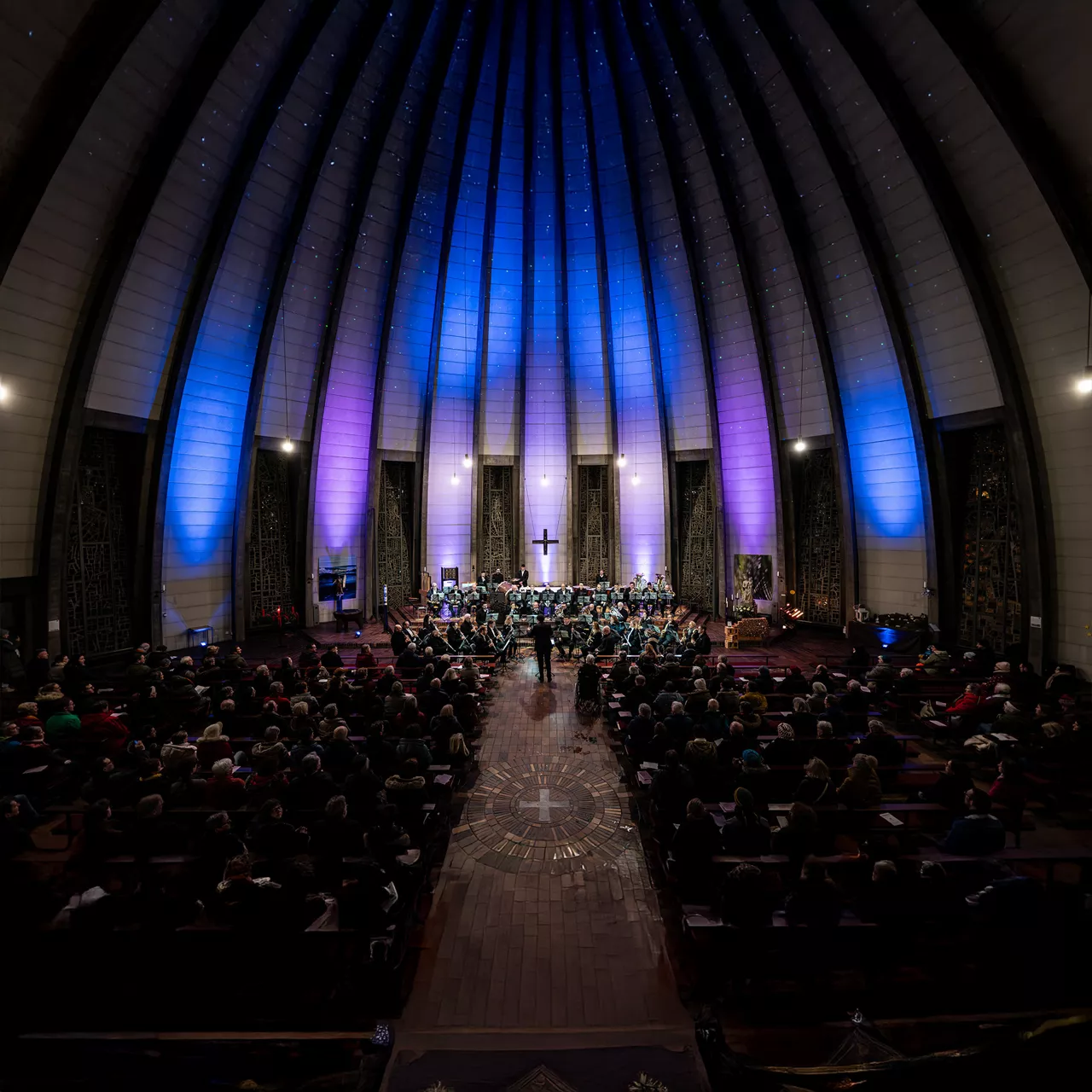 Blick in eine Kirche mit hoher, beleuchteter Decke, ein Orchester spielt vor einem sitzenden Publikum.