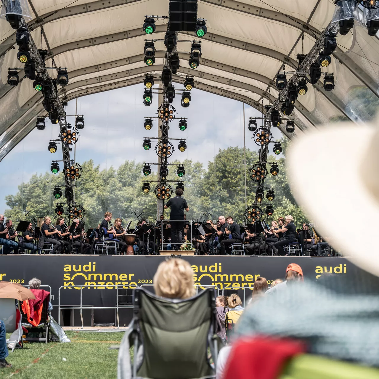 Open-Air-Bühne mit Orchester unter einem Zeltdach, Publikum sitzt auf dem Rasen vor der Bühne.
