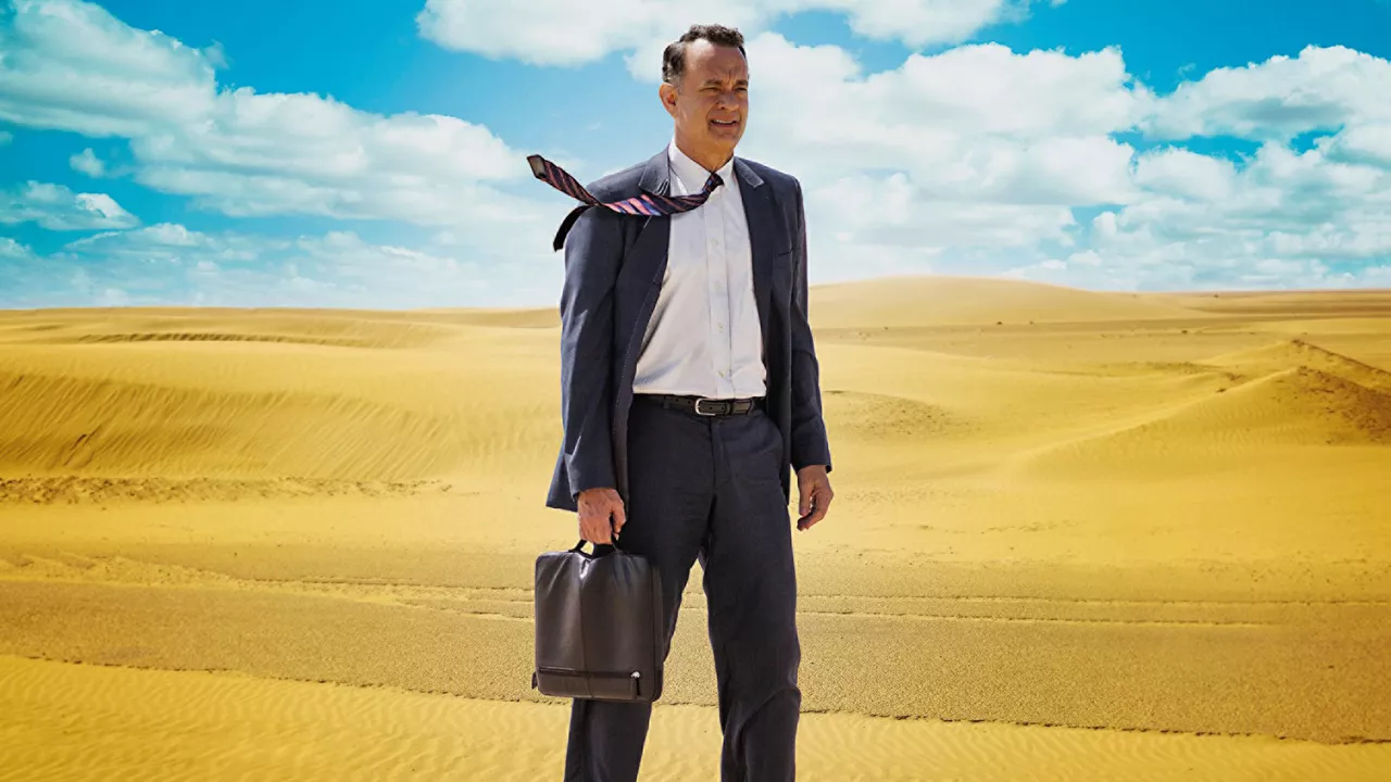 Mann im Anzug (Tom Hanks) mit Koffer in der Hand steht in einer Wüste bei blauem Himmel mit leichten Wolken. Der Schlips weht nach links zur Seite.