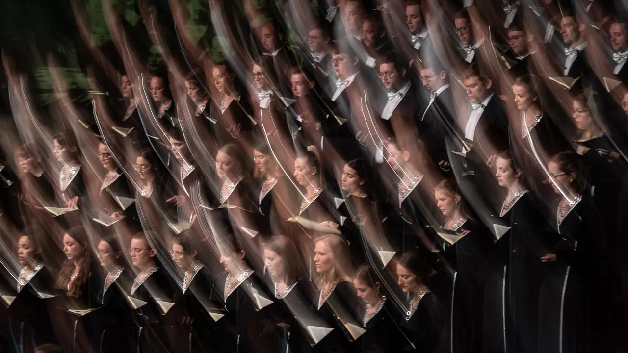 Chor in schwarzen Konzertoutfits singt mit Noten in der Hand; künstlerisch verschwommene Bewegungsunschärfe erzeugt dynamischen Effekt.