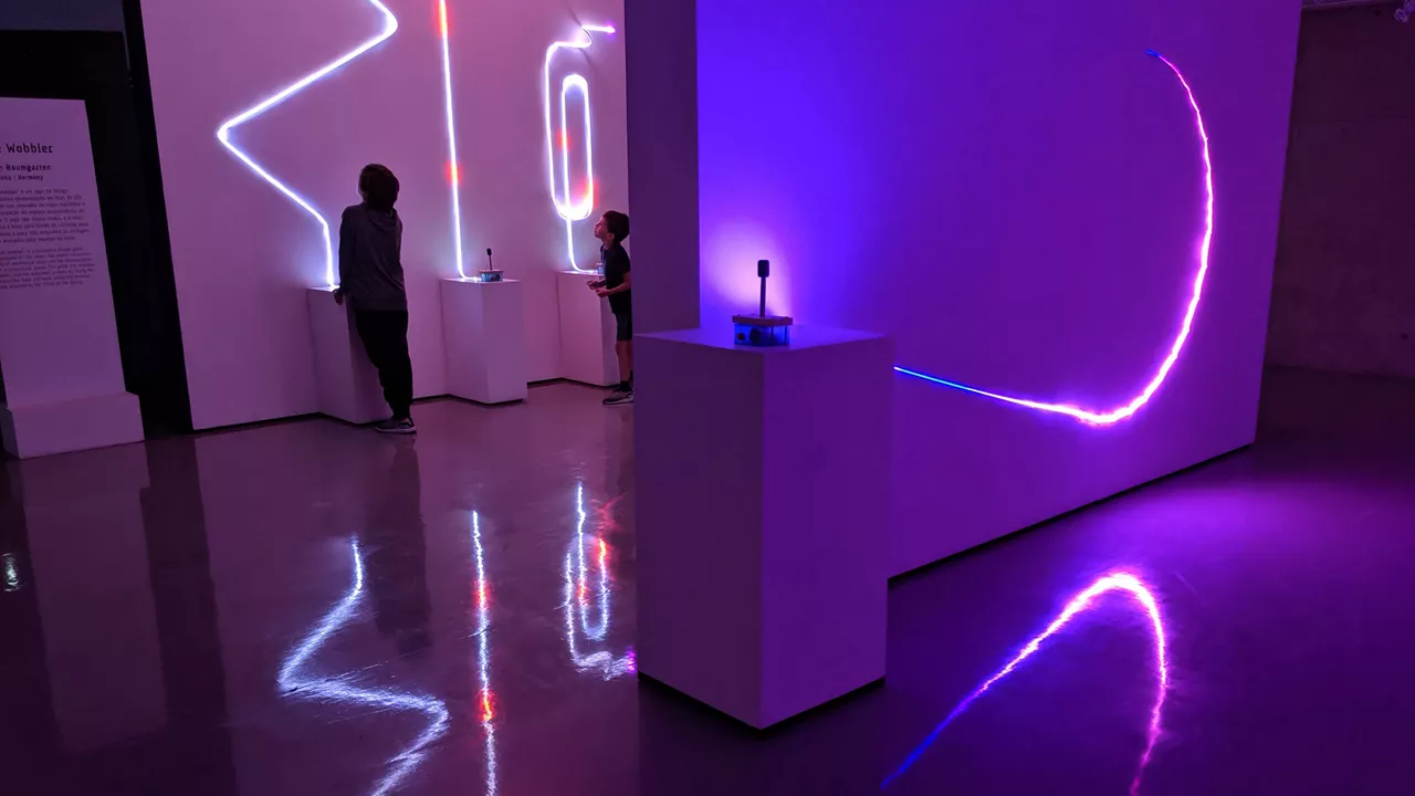 Zwei Personen betrachten leuchtende bunte LED-Linien an weißen Wänden in dunklem Ausstellungsraum.