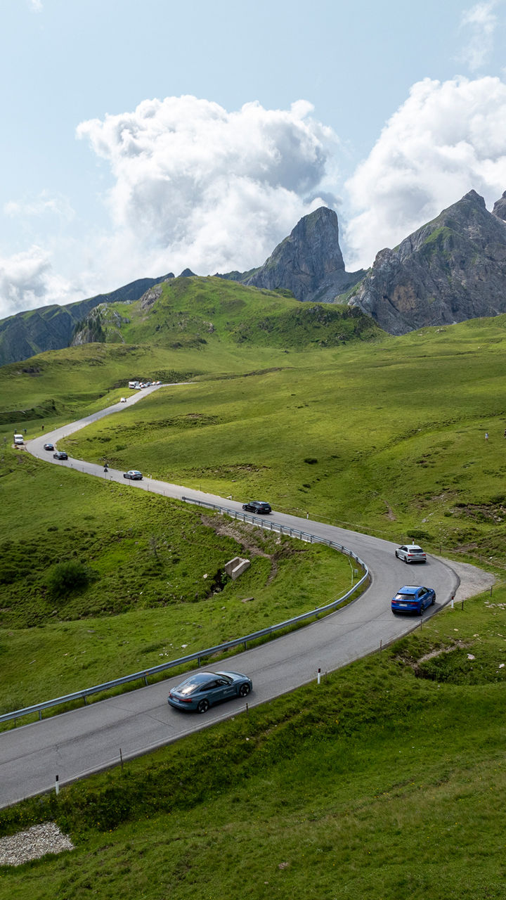 Vogelperspektive einer Landschaft mit einer kurvigen Straße, auf der hintereinander mehrere Audi Modelle fahren, im Hintergrund befinden sich Berge