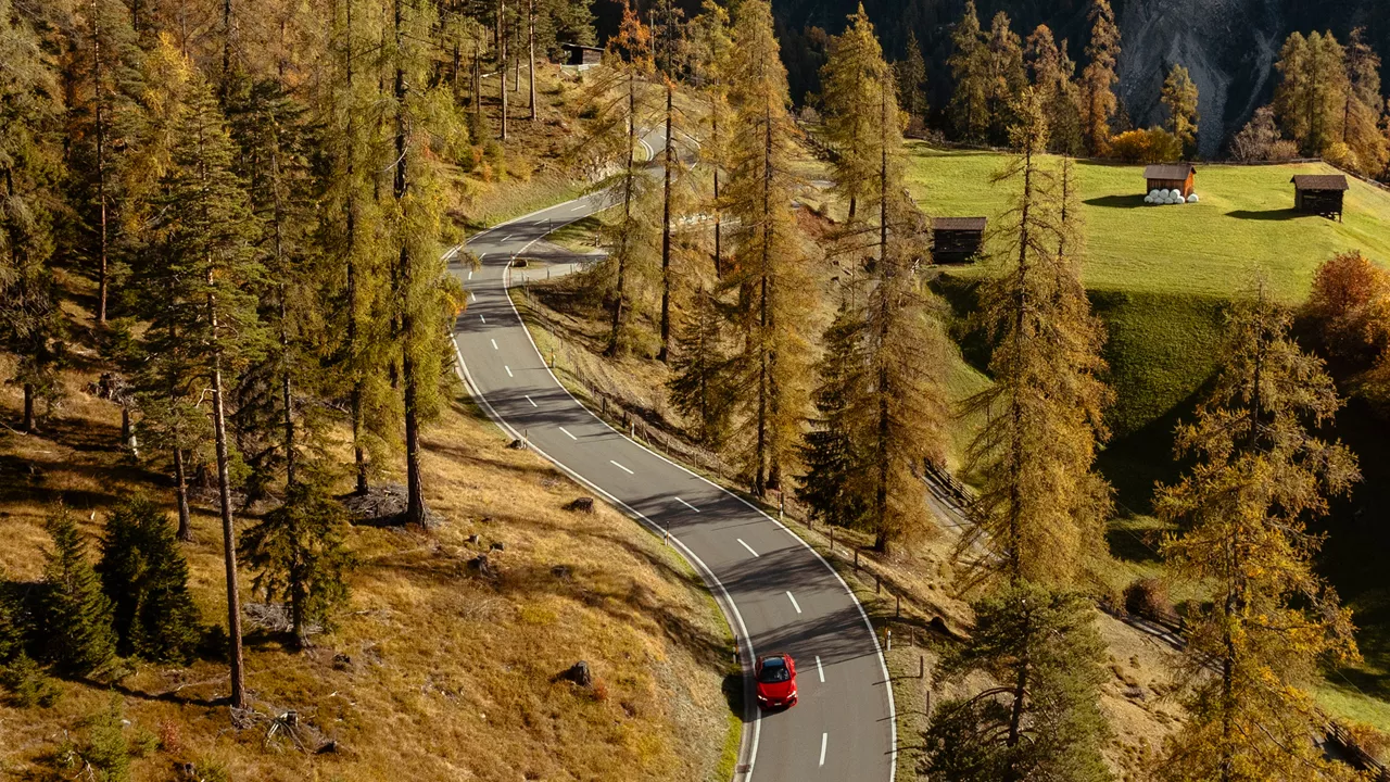 Red Audi model with panoramic roof drives on a winding road through an autumnal mountain landscape with scattered wooden huts.