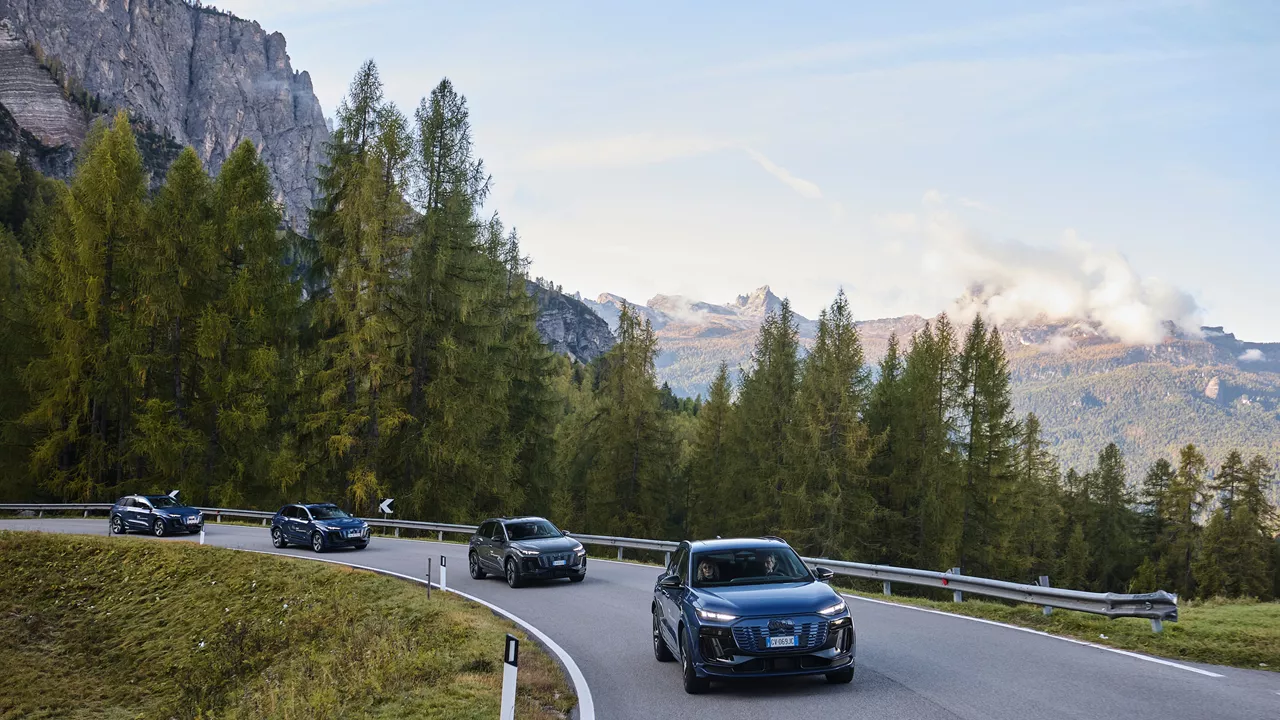 Four dark blue and black Audi SUV models drive through a wooded mountain bend with a rocky background and blue sky.