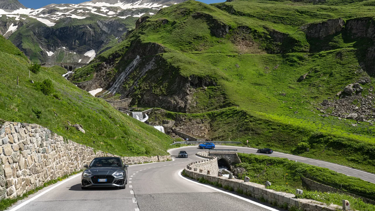 Audi models driving on an alpine road with a green slope, stone wall and waterfall in the background.