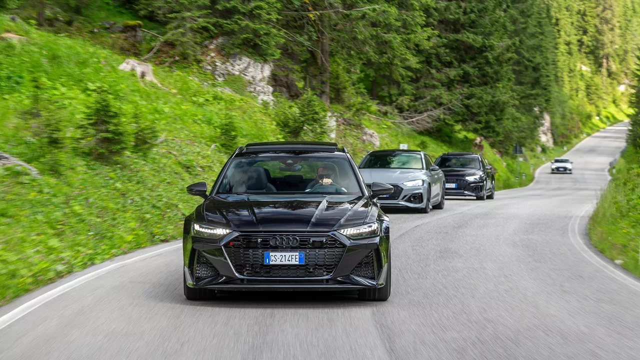 Black Audi RS model leads convoy of vehicles through wooded mountain road with winding route.