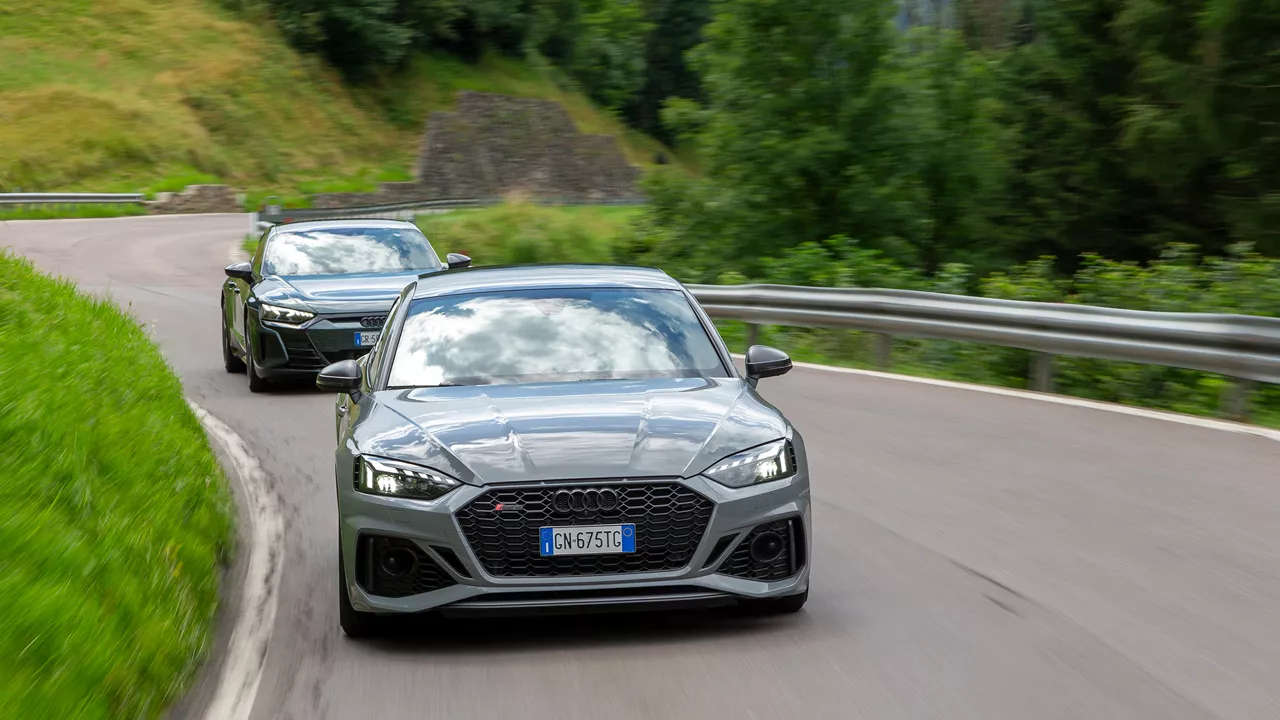 Two Audi sports cars in gray and dark green drive closely behind each other through a sharp bend on a hilly country road.