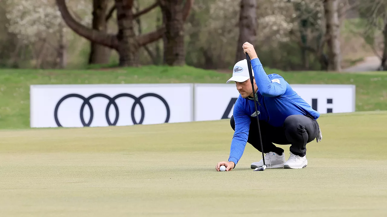 Man in blue training jacket with white cap squats on golf course with Audi logo in the background and concentrates on the putt.