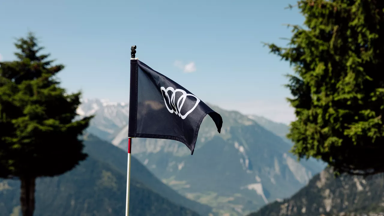 Black flag with white Audi logo fluttering on golf course, mountains and conifers in the background under a blue sky.