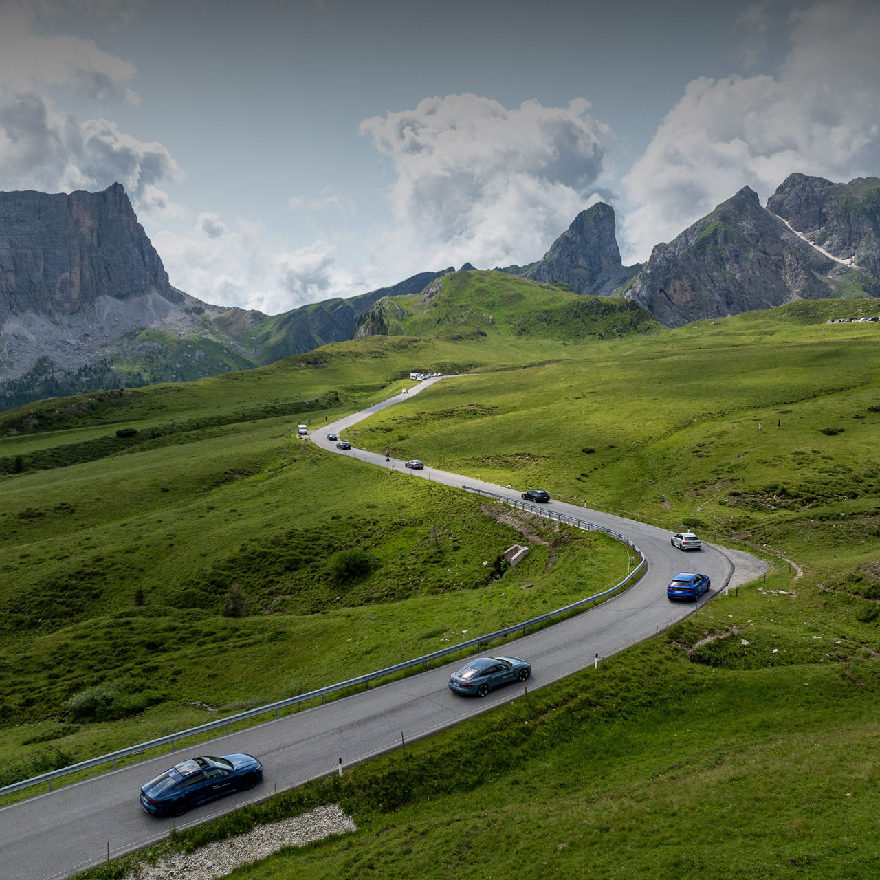 Vogelperspektive einer Landschaft mit einer kurvigen Straße, auf der hintereinander mehrere Audi Modelle fahren, im Hintergrund befinden sich Berge. Zu den Events.