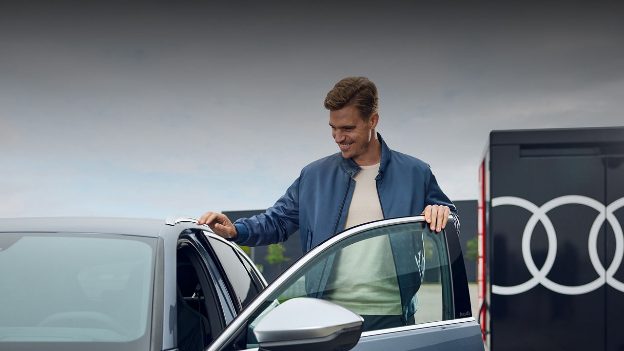 Mann steht vor einer geöffneten Autotür eines Audi Modells, im Hintergrund ist ein Audi Logo auf einem Gebäude zu sehen. Zur My Audi driving experience erleben. 