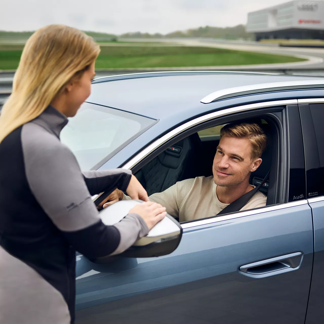 A woman with blond hair (rear view) explains something to a man in a car, she stands in front of the car with documents and the man has his window sticker down 