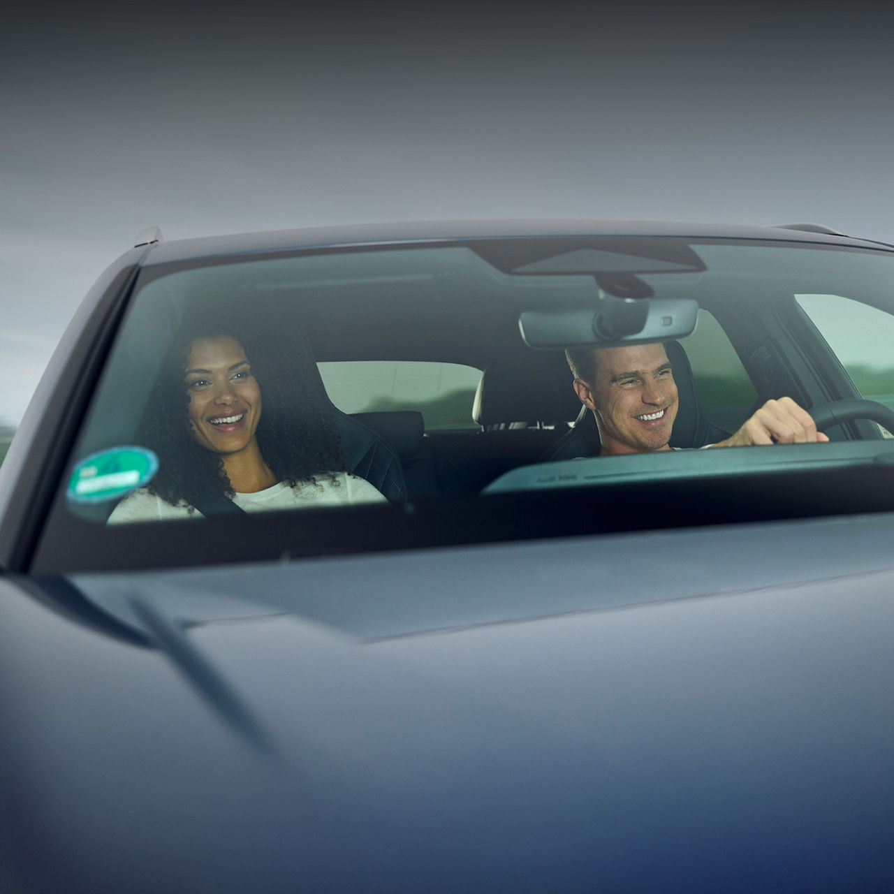 Zwei Personen im Auto, Fahrer lächelt während der Fahrt, Innenansicht durch die Frontscheibe.