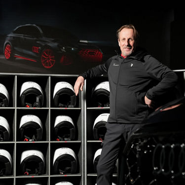 Instructor Frank Biela leans casually against a rack of helmets. He is wearing a black sweatshirt with a white Audi logo. 