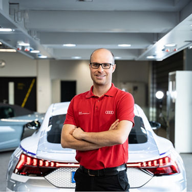 Instructor Nils Mierschke wears a red Audi polo shirt. With his arms folded, he stands in front of the rear of an Audi model and looks into the camera with a smile. 