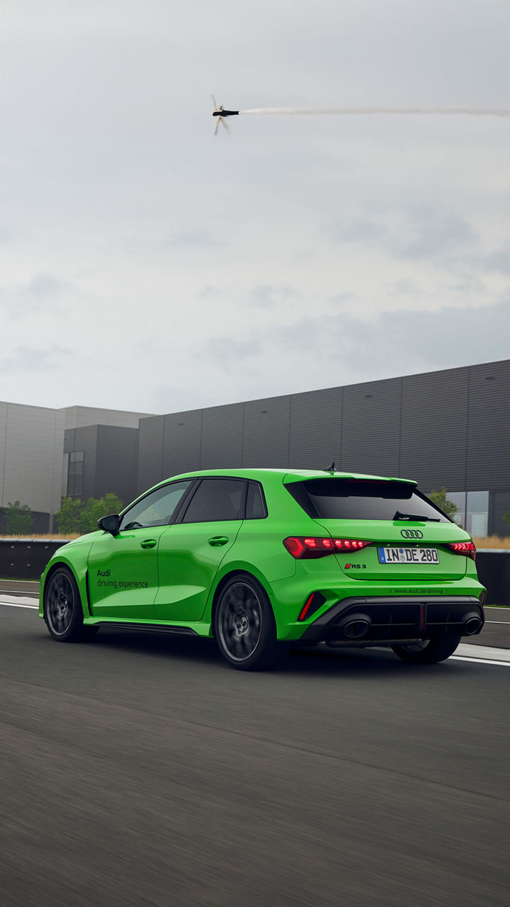 A green Audi RS 3 Sportback drives past the Audi driving experience Center on a race track.