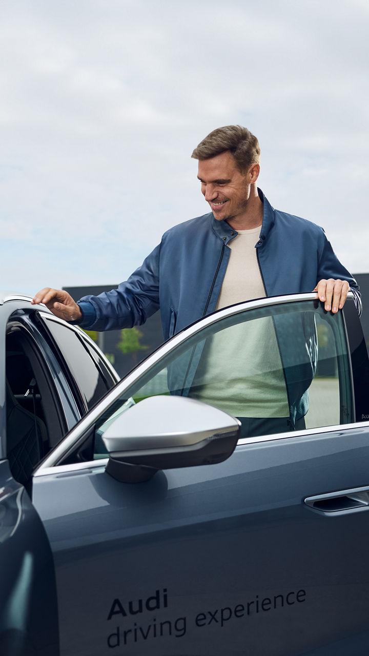 Mann steht vor einer geöffneten Autotür eines Audi Modells, im HIntergrund sieht man ein Audi Logo auf einem Gebäude