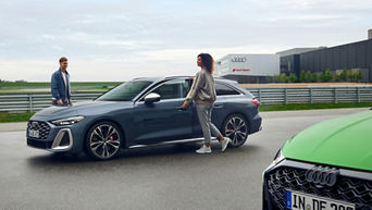 A man and a woman in sporty clothing get into an Audi model. An Audi driving centre building can be seen in the background.