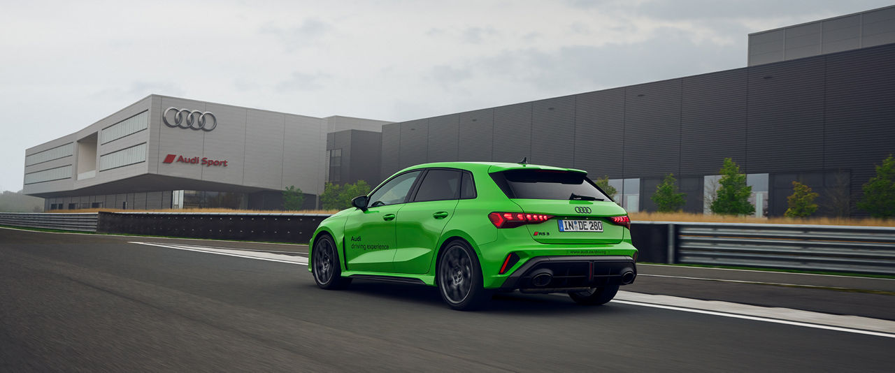 Hellgrüner Audi RS 3 Sportback fährt auf einer Strecke in Neuburg, im Hintergrund ist ein großes Gebäude mit Audi Sport Logo