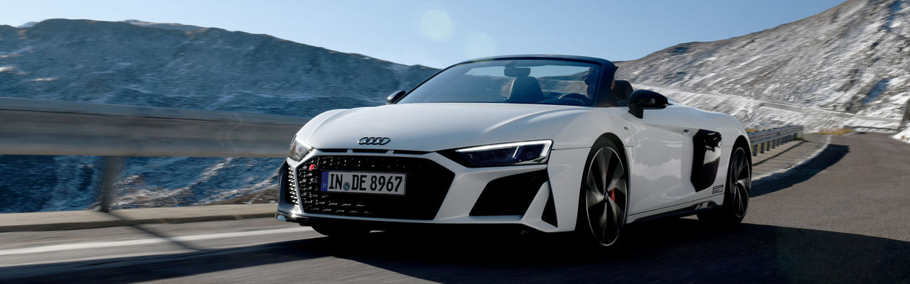 White Audi R8 Spyder driving on a road through a mountainous landscape with light snow on the mountains