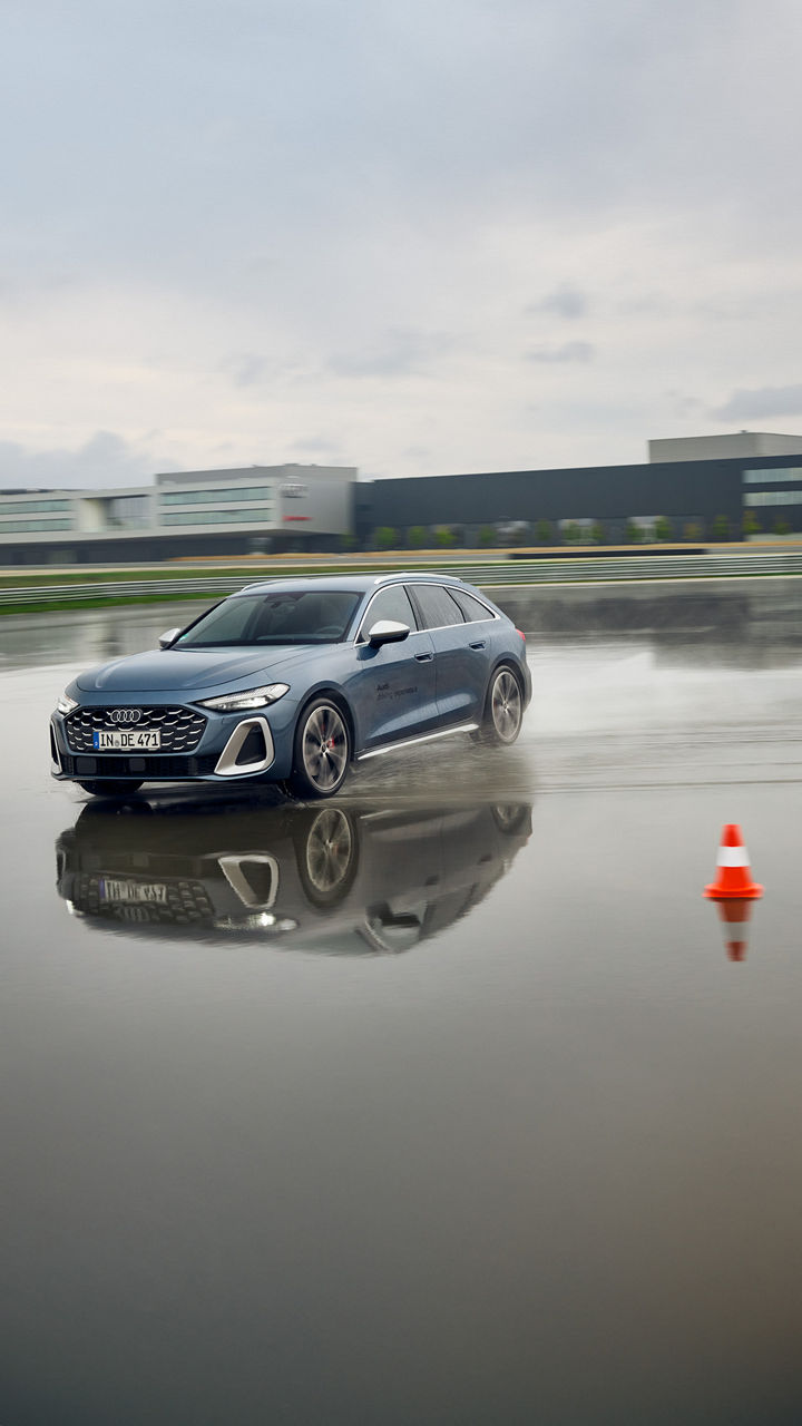 Audi S5 Avant fährt auf einem nassen Übungsgelände durch eine tiefe Pfütze, im Hintergrund befindet sich ein Audi Gebäude