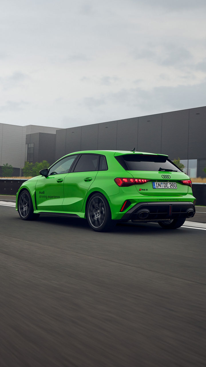 Light green Audi RS 3 Sportback on a training ground, in the background is a building with the Audi Sport logo