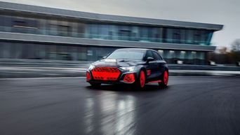Audi RS 3 driving experience fährt auf einer Übungsstecke bei grauem Himmel in der Dämmerung, im Hintergrund befindet sich ein großes Gebäude mit Glasfronten.