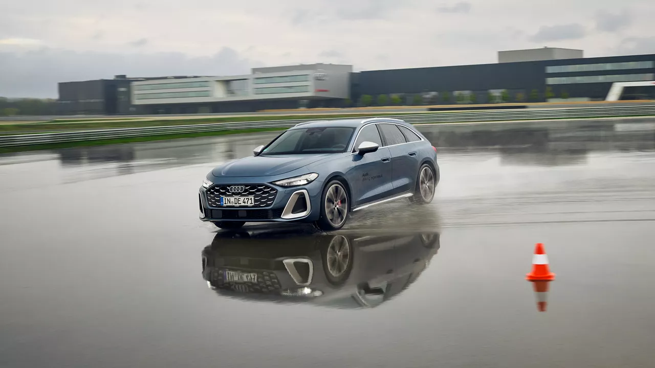 Audi S5 Avant drives through a deep puddle on a wet training ground, with an Audi building in the background