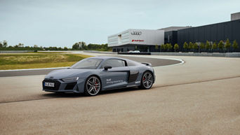 Mattgrauer Audi R8 Coupé mit weißer Audi driving experience Aufschrift auf Fahrertür fährt auf einer Strecke, im Hintergrund befindet sich ein Audi Sport Gebäude.