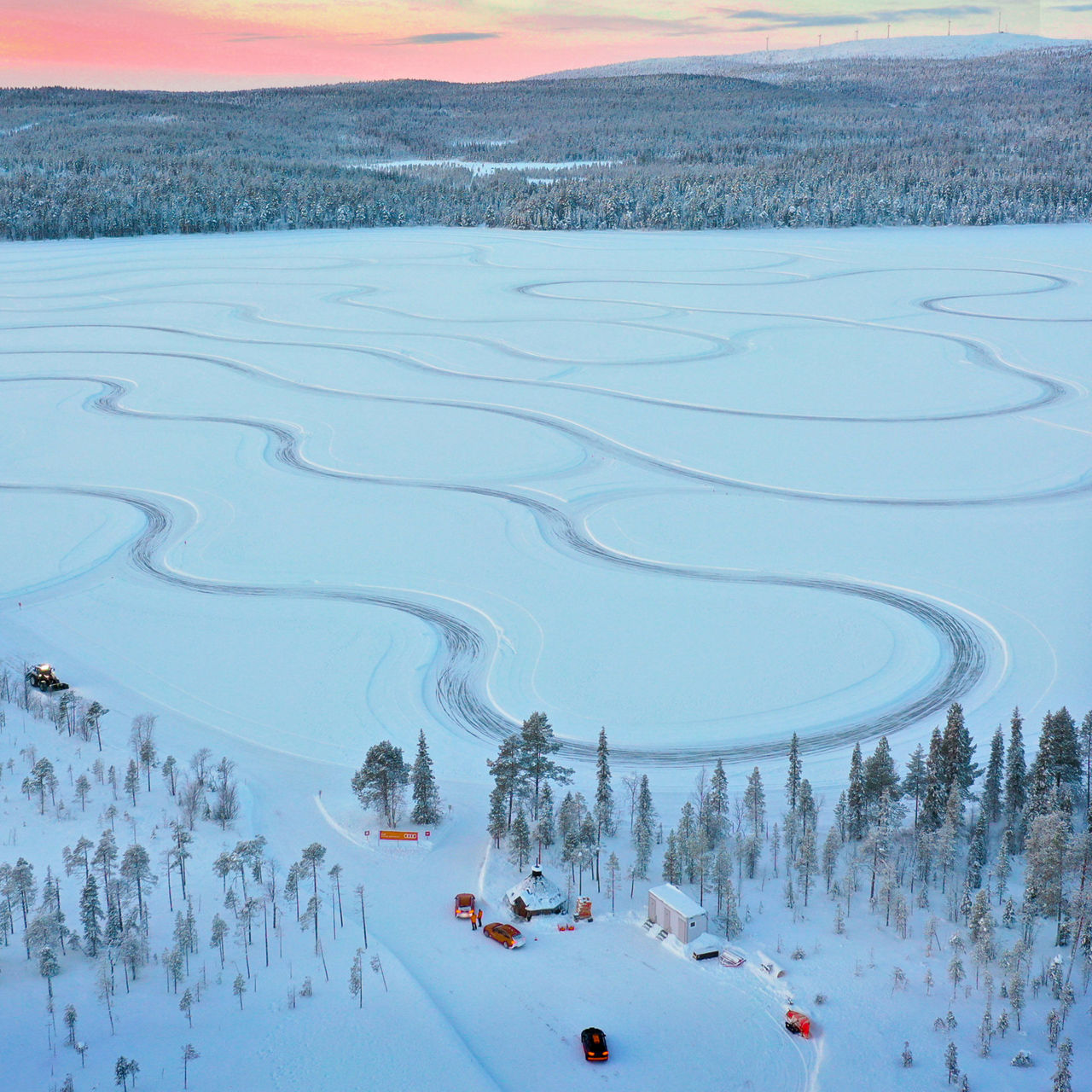 Bird's eye view of an icy surface and a forest with a pink, yellowish sky and vehicle tracks