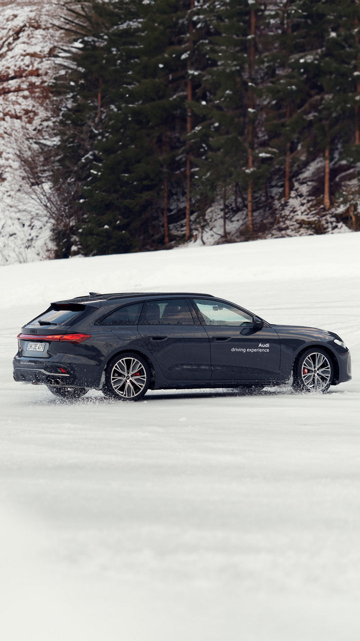 Dunkler Audi S5 Avant mit weißem Schriftzug „Audi driving experience“ driftet über schneebedeckte Fläche.