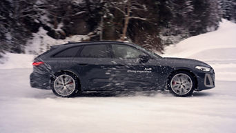 Dark Audi S5 Avant with white “Audi driving experience” lettering driving on a snow-covered road, snow swirled up in the focus.