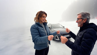 Two people (a younger woman and an older man with glasses) in winter clothing stand on a patch of ice and simulate driving movements, while an Audi can be seen in the background.