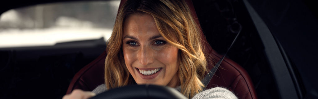 Nahaufname Frau mit blonden Haaren lächelnd beim Autofahren, Sicherheitsgurt sichtbar.