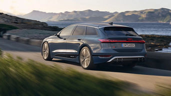 Grauer Audi S6 Avant e-tron fährt auf einer Küstenstraße entlang des Meeres, im Hintergrund Berge und leicht bewölkter Himmel.