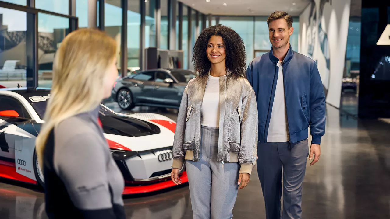 Two smiling people talking to an employee in an Audi showroom with a sports car in the background.