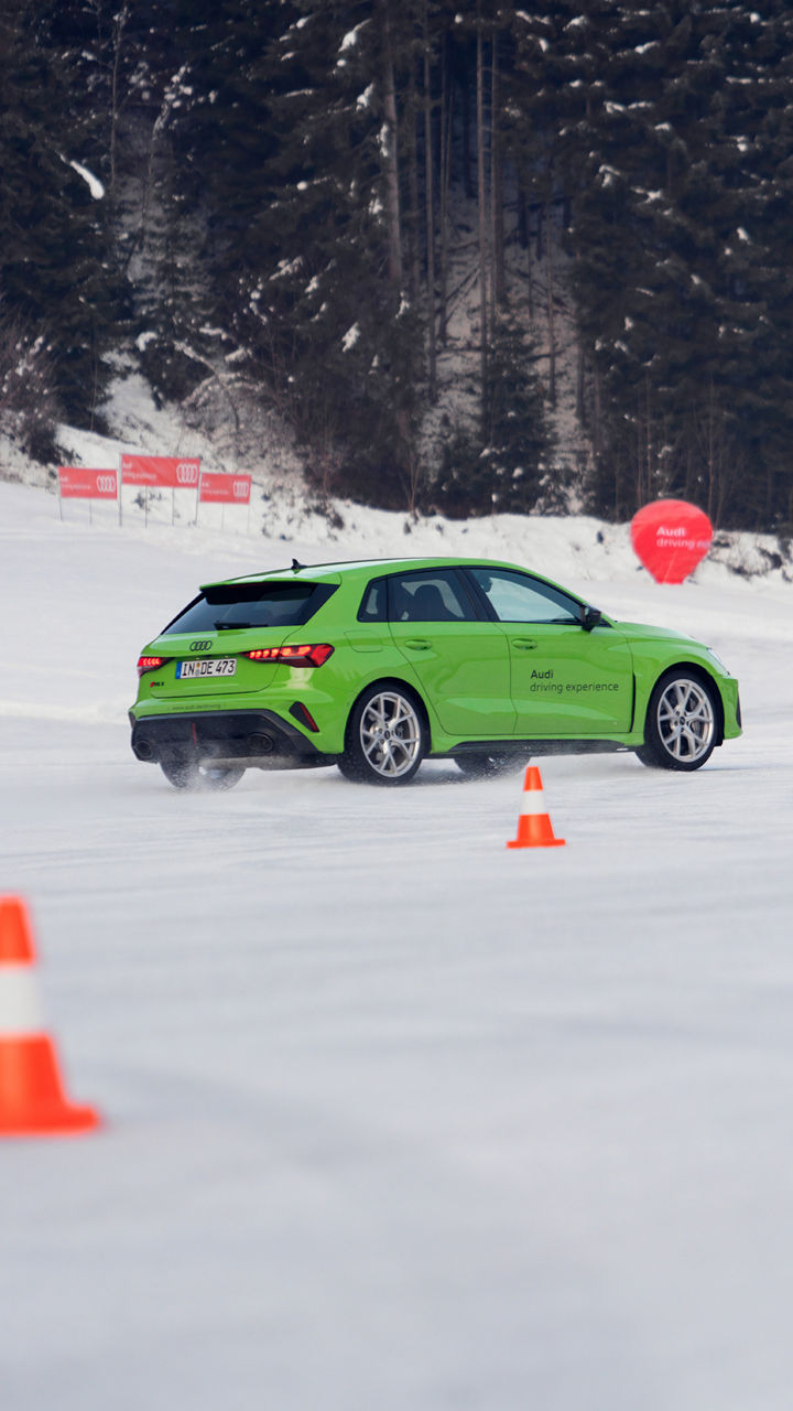 Hellgrüner Audi RS 3 Sportback fährt auf schneebedeckter Strecke zwischen orangen Pylonen, im Hintergrund verschneiter Wald.