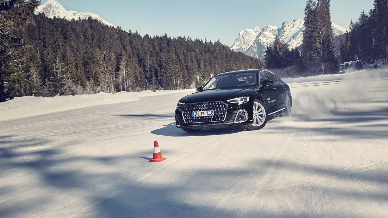 Schwarzer Audi A8 50 TDI quattro fährt im Schnee durch Parcour, im Hintergrund blauer Himmel, Wälder und Berge