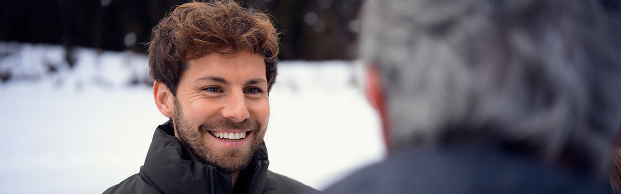 Smiling man with brown curls in a winter outfit talking to a gray-haired man in the foreground, snowy landscape in the background.