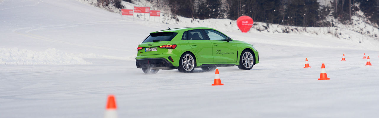 Light green Audi RS 3 Sportback driving on a snow-covered track between orange cones, snow-covered forest in the background.