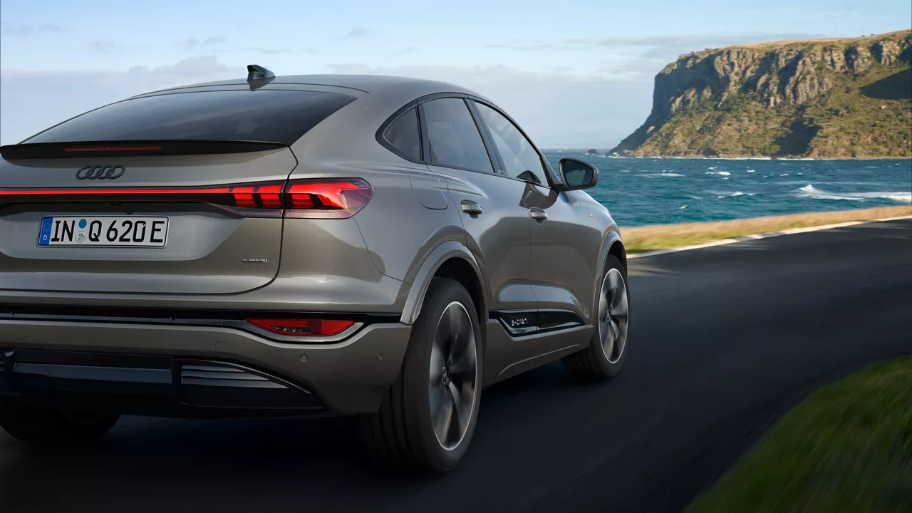 Grauer Audi Q6 Sportback e-tron fährt an Küstenstraße entlang, Blick auf das Heck des Fahrzeugs mit Meer und Klippen im Hintergrund.