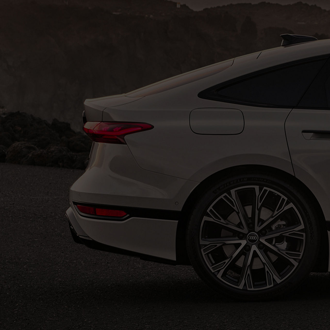 Heckansicht eines weißen Audi mit Rücklicht und Felge, im Hintergrund eine Felslandschaft bei Abendstimmung.