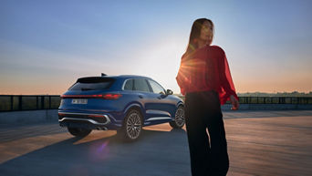 Frau im roten Oberteil steht neben einem blauen Audi SUV bei Sonnenuntergang auf einem Parkdeck mit weitem Ausblick.