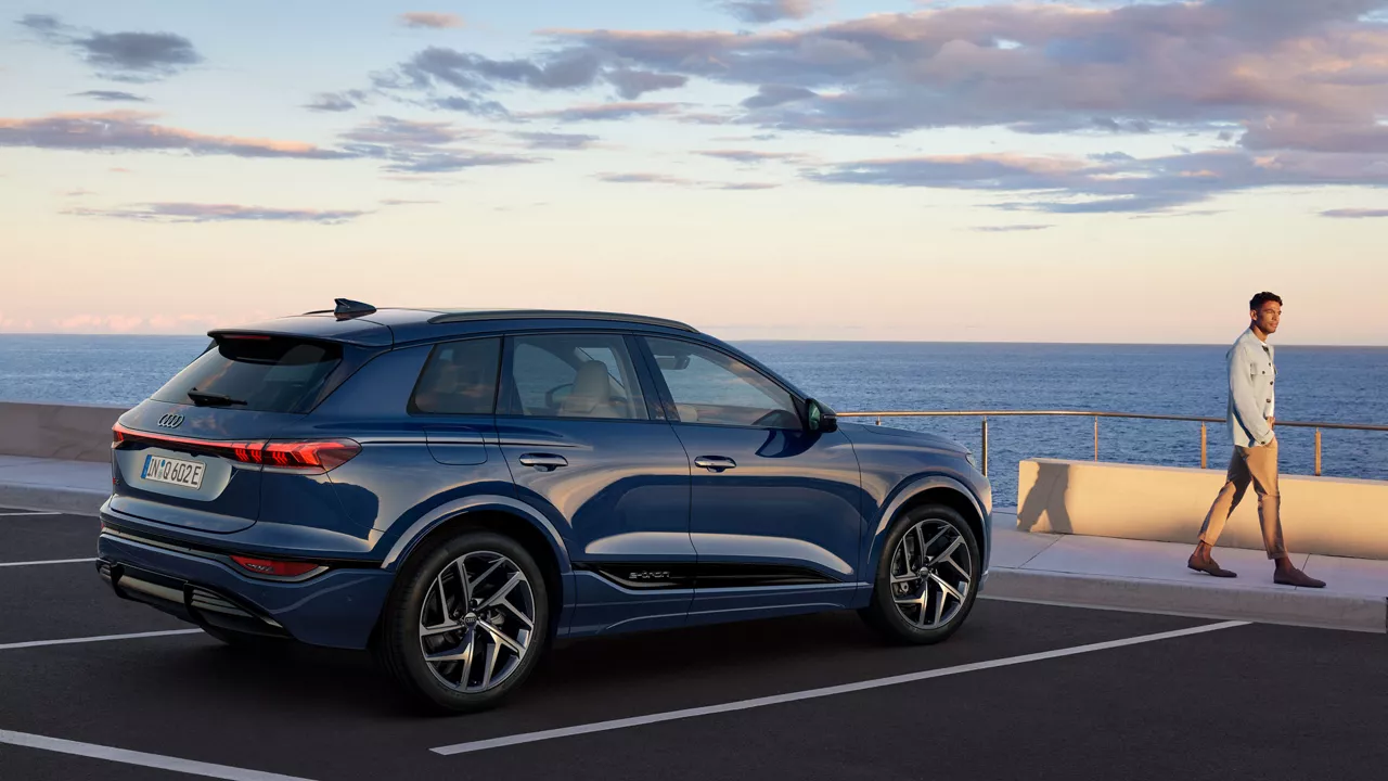 Blauer Audi-SUV steht auf einem Parkplatz am Meer, ein Mann in heller Kleidung geht am Geländer entlang.