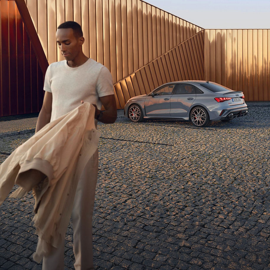 Ein Mann läuft von einem silbernen Audi weg, schaut auf den Boden und trägt eine Jacke in einer Hand
