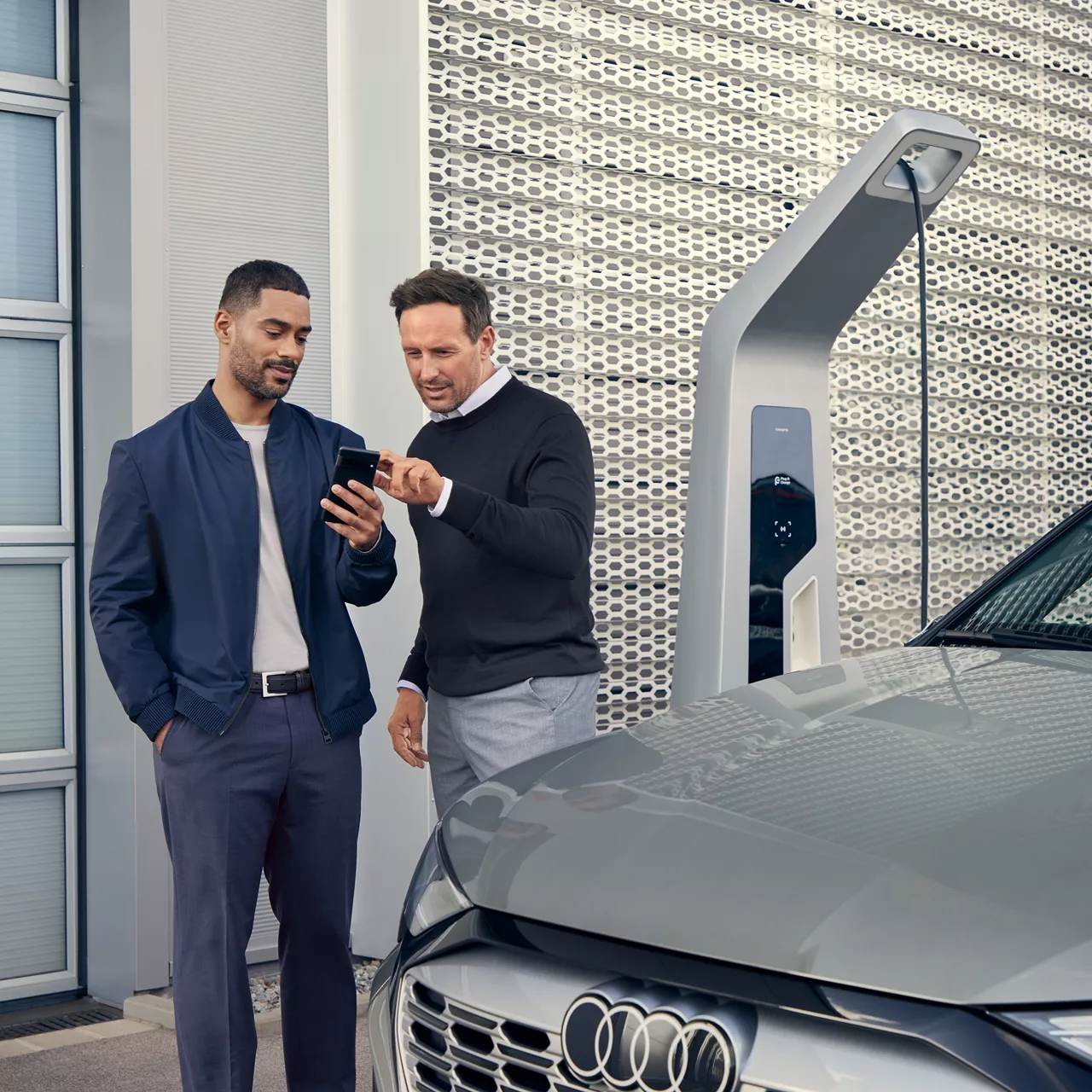 Ein Mann zeigt einem anderen Mann etwas auf dem Handy, während sie einen Audi in einer Ladestation vor sich haben