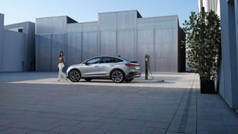 Eine stilvoll gekleidete Frau geht auf einem modernen, weitläufigen Platz an einem Audi Q4 e-tron vorbei, der an einer Ladestation angeschlossen ist. Im Hintergrund sind moderne, futuristische Gebäude zu sehen.