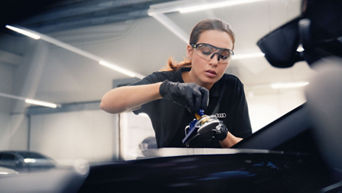 Mechanikerin mit Schutzbrille behandelt die Frontscheibe mit elektrischem Werkzeug in Werkstatt.