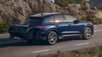 Blauer Audi Q4 e-tron fährt mit großem schwarzen Heckgepäckträger auf Landstraße entlang felsiger Landschaft.