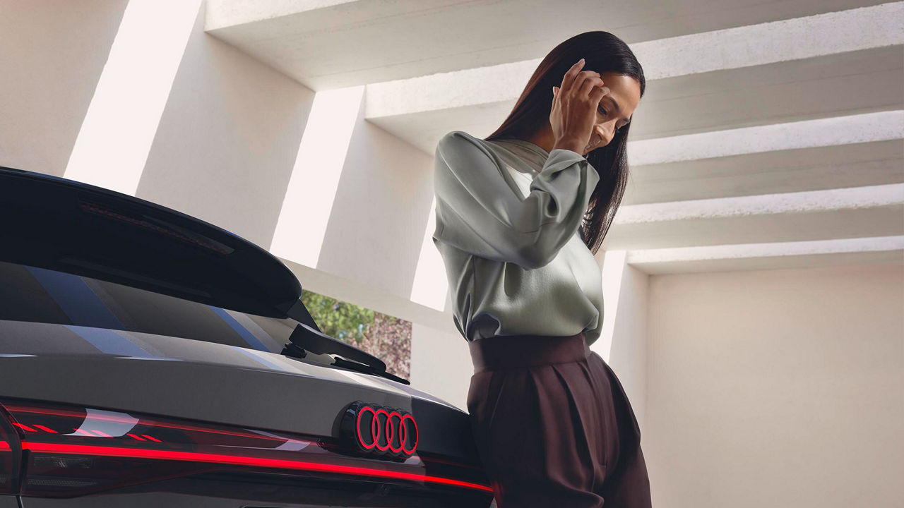 Frau lehnt an einem grauen Audi mit leuchtenden Rücklichtern, Innenhof mit Lichtschächten im Hintergrund.
